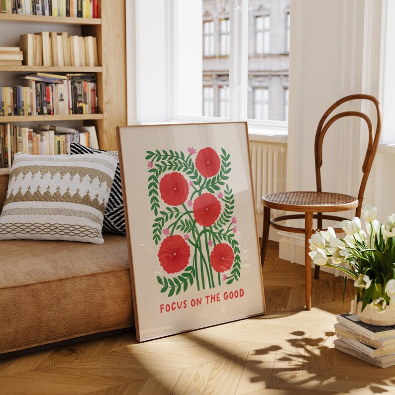May include: A framed art print featuring a floral design with red flowers and green leaves. The print includes the text "FOCUS ON THE GOOD" at the bottom. The artwork is displayed in a well-lit room with a wooden floor and a bookshelf.