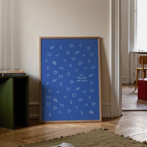 May include: A framed blue print with white line drawings of various facial expressions and the text "ALL FEELINGS ARE OKAY". The print is in a wooden frame and is displayed against a neutral wall.