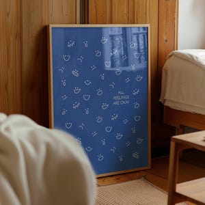 May include: A framed blue poster featuring white line drawings of different facial expressions. The text "ALL FEELINGS ARE OKAY" is printed in white. The poster is in a wooden frame and is leaning against a wooden wall.