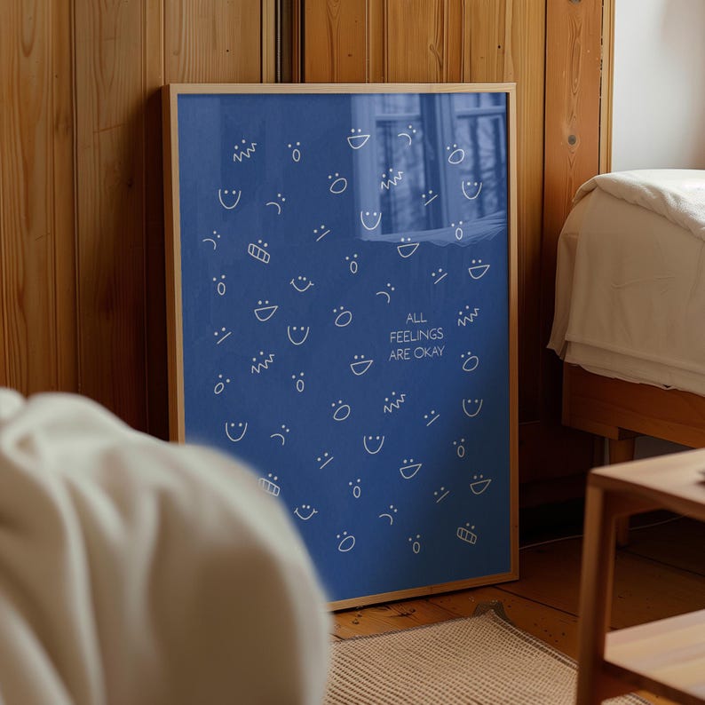May include: A framed blue poster featuring white line drawings of different facial expressions. The text "ALL FEELINGS ARE OKAY" is printed in white. The poster is in a wooden frame and is leaning against a wooden wall.