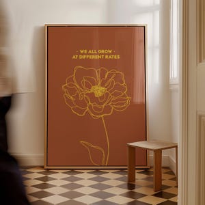 Puede incluir: Un cuadro enmarcado con un contorno floral amarillo sobre un fondo color óxido. El texto "WE ALL GROW - AT DIFFERENT RATES" está encima de la flor. El marco es dorado. Una pequeña mesa auxiliar de madera está cerca.