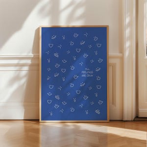 May include: A framed art print featuring a blue background adorned with white, stylized faces and shapes. The text "ALL FEELINGS ARE OKAY" is centered. The frame is a light brown colour.