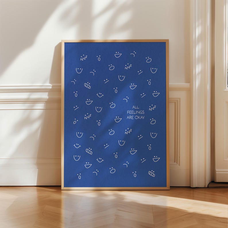 May include: A framed art print featuring a blue background adorned with white, stylized faces and shapes. The text "ALL FEELINGS ARE OKAY" is centered. The frame is a light brown colour.
