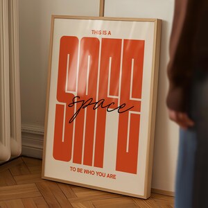 May include: A framed poster with the words "THIS IS A GOOD SPACE" in large, orange block letters. The word "space" is written in cursive. The phrase "TO BE WHO YOU ARE" is at the bottom. The poster is in a light wood frame.