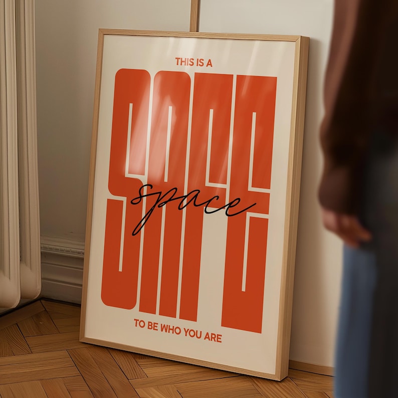 May include: A framed poster with the words "THIS IS A GOOD SPACE" in large, orange block letters. The word "space" is written in cursive. The phrase "TO BE WHO YOU ARE" is at the bottom. The poster is in a light wood frame.