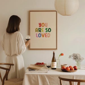 May include: A framed print with the text "YOU ARE SO LOVED" in colorful letters. The print is hanging on a white wall above a table set with wine glasses, a bottle of wine, and fruit.