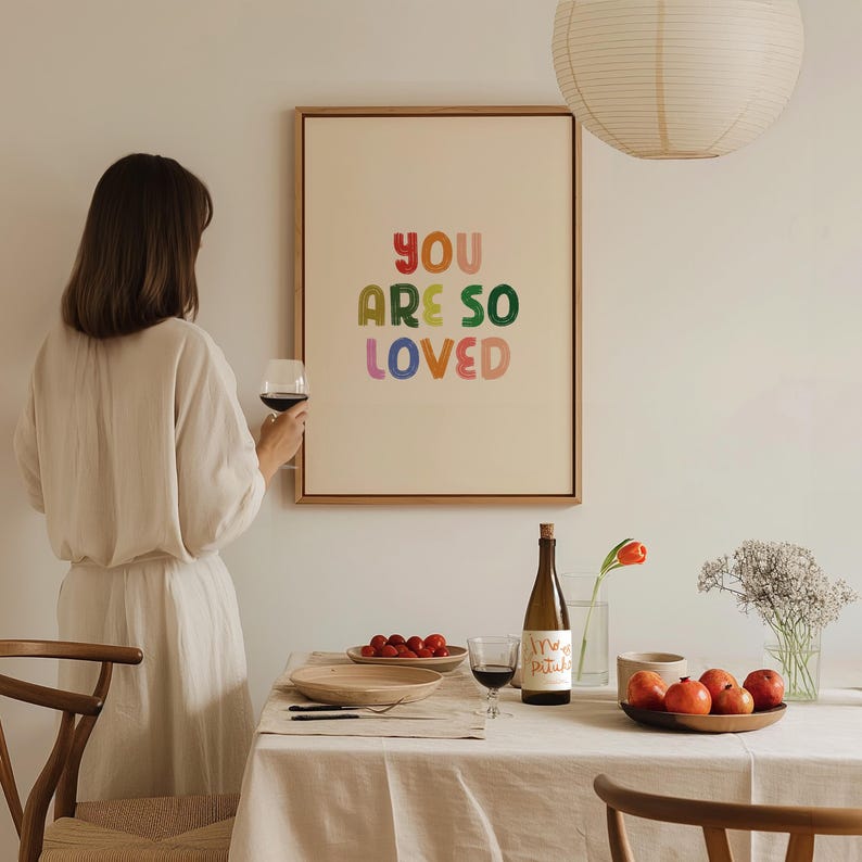 May include: A framed print with the text "YOU ARE SO LOVED" in colorful letters. The print is hanging on a white wall above a table set with wine glasses, a bottle of wine, and fruit.