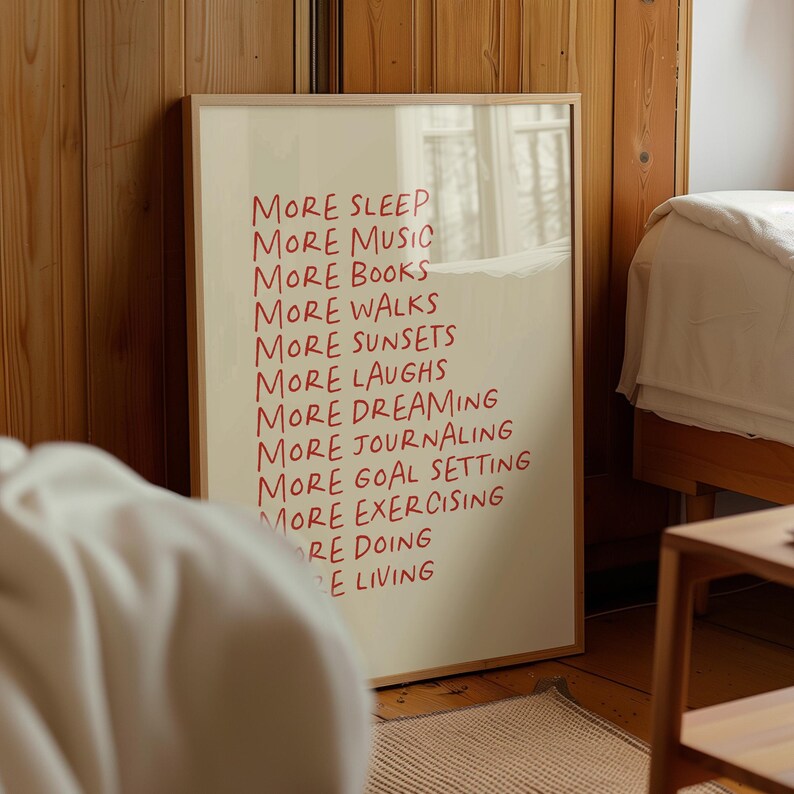 May include: A beige framed print with red text that reads "More Sleep, More Music, More Books, More Walks, More Sunsets, More Dreaming, More Laughs, More Journaling, More Goal Setting, More Exercising, More Doing, More Living."