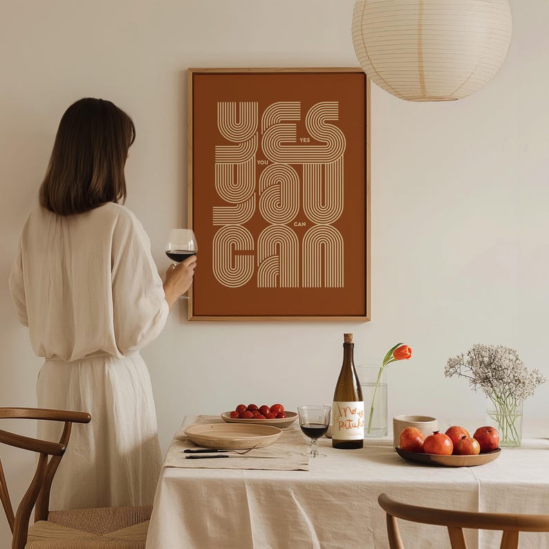 Pu&ograve; includere: Una stampa incorniciata marrone con il testo "YES YOU CAN" in uno stile di font retr&ograve;. La stampa &egrave; appesa a un muro bianco sopra un tavolo apparecchiato con una bottiglia di vino, bicchieri da vino e frutta.