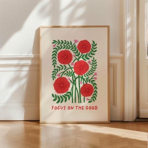May include: A framed art print featuring a floral design with red flowers, green leaves, and pink accents. The print includes the text "FOCUS ON THE GOOD" in a bold font. The artwork is displayed in a light-colored wooden frame.