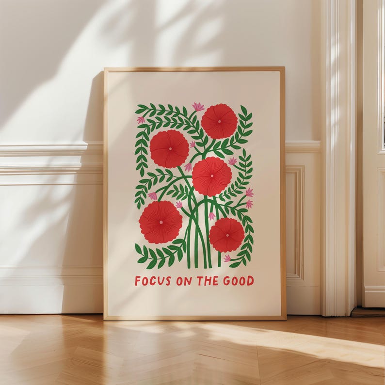 May include: A framed art print featuring a floral design with red flowers, green leaves, and pink accents. The print includes the text "FOCUS ON THE GOOD" in a bold font. The artwork is displayed in a light-colored wooden frame.