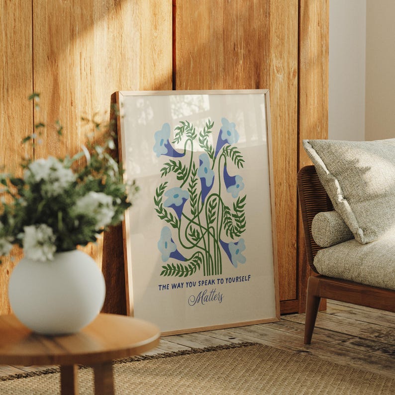 May include: A framed art print featuring a floral design with blue bell-shaped flowers and green leaves. The text "THE WAY YOU SPEAK TO YOURSELF Matters" is below the illustration. The artwork is displayed in a room with a wooden floor and furniture.