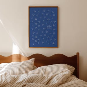 May include: A framed blue print with white line drawings of various faces and shapes. The text "ALL FEELINGS ARE OKAY" is centered on the print. The print is in a brown wooden frame and hangs above a bed with striped bedding.