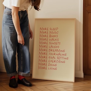May include: A framed print with the text "More Sleep, More Music, More Books, More Walks, More Sunsets, More Laughs, More Dreaming, More Journaling, More Goal Setting, More Exercising, More Doing, More Living" written in red on a cream background.
