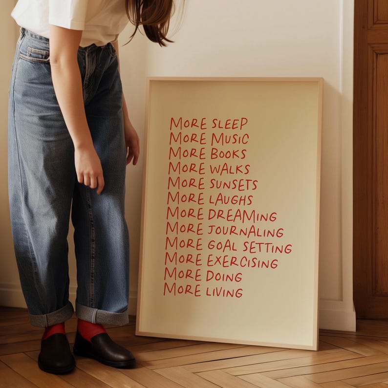 May include: A framed print with the text "More Sleep, More Music, More Books, More Walks, More Sunsets, More Laughs, More Dreaming, More Journaling, More Goal Setting, More Exercising, More Doing, More Living" written in red on a cream background.
