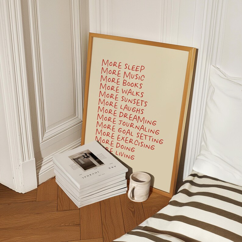 May include: A framed print with a beige background and red text that reads "More Sleep, More Music, More Books, More Walks, More Sunsets, More Laughs, More Dreaming, More Journaling, More Goal Setting, More Exercising, More Doing, More Living".
