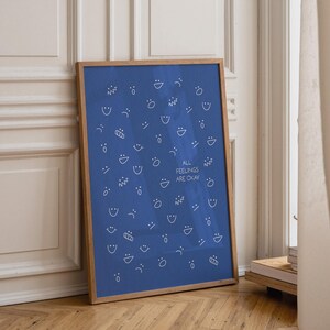 May include: A framed poster with a blue background and white line drawings of various facial expressions. The text "ALL FEELINGS ARE OKAY" is centered on the poster. The frame is made of light brown wood.