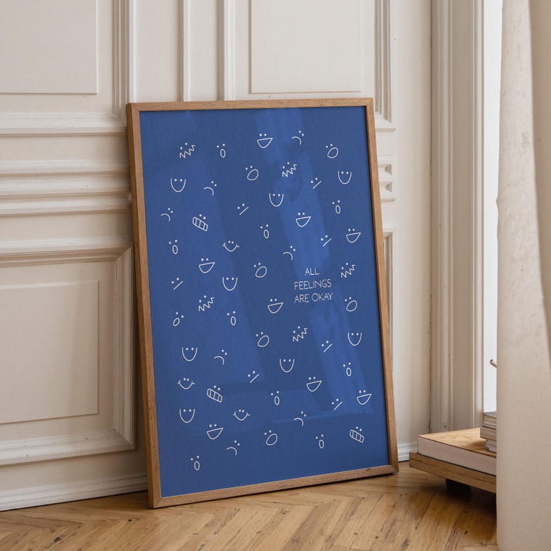 May include: A framed poster with a blue background and white line drawings of various facial expressions. The text "ALL FEELINGS ARE OKAY" is centered on the poster. The frame is made of light brown wood.