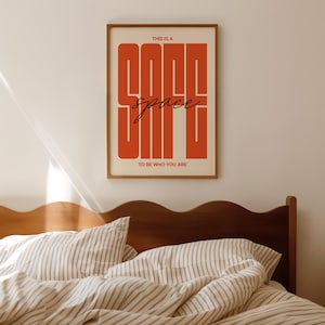 May include: A framed art print with the words "THIS IS A GOOD space TO BE WHO YOU ARE" in a bold, orange font. The print is in a wooden frame and hangs above a bed with striped bedding.