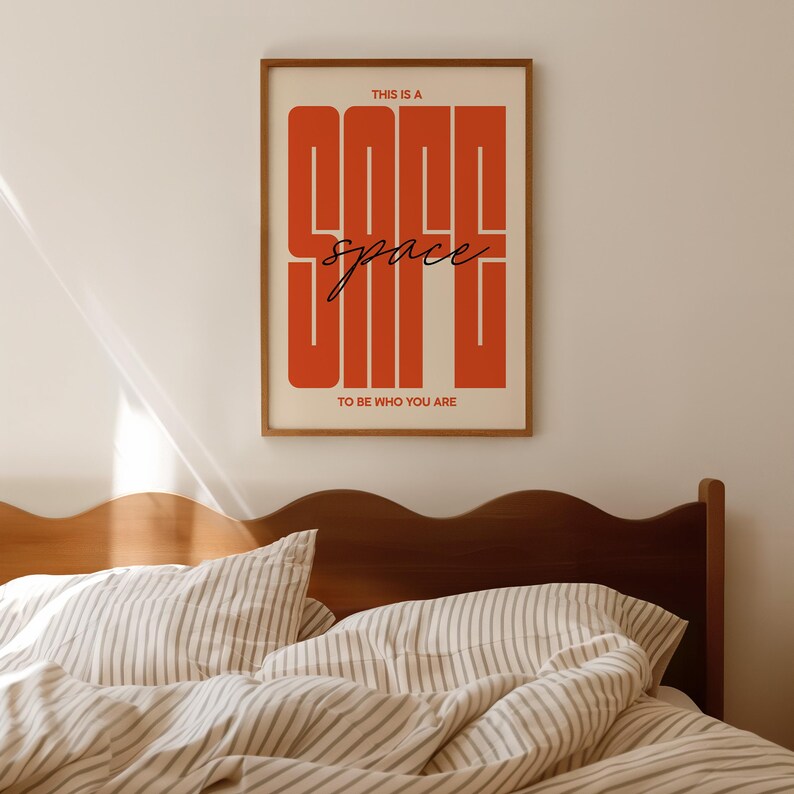 May include: A framed art print with the words "THIS IS A GOOD space TO BE WHO YOU ARE" in a bold, orange font. The print is in a wooden frame and hangs above a bed with striped bedding.