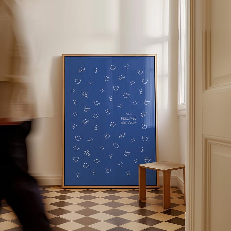 May include: A framed blue art print with a gold frame. The print features white line drawings of various facial expressions and the text "ALL FEELINGS ARE OKAY". A small wooden stool is next to it.
