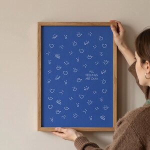 May include: A framed blue print featuring a pattern of white line-drawn faces and shapes. The print includes the text "ALL FEELINGS ARE OKAY" in white. The frame is made of light brown wood.