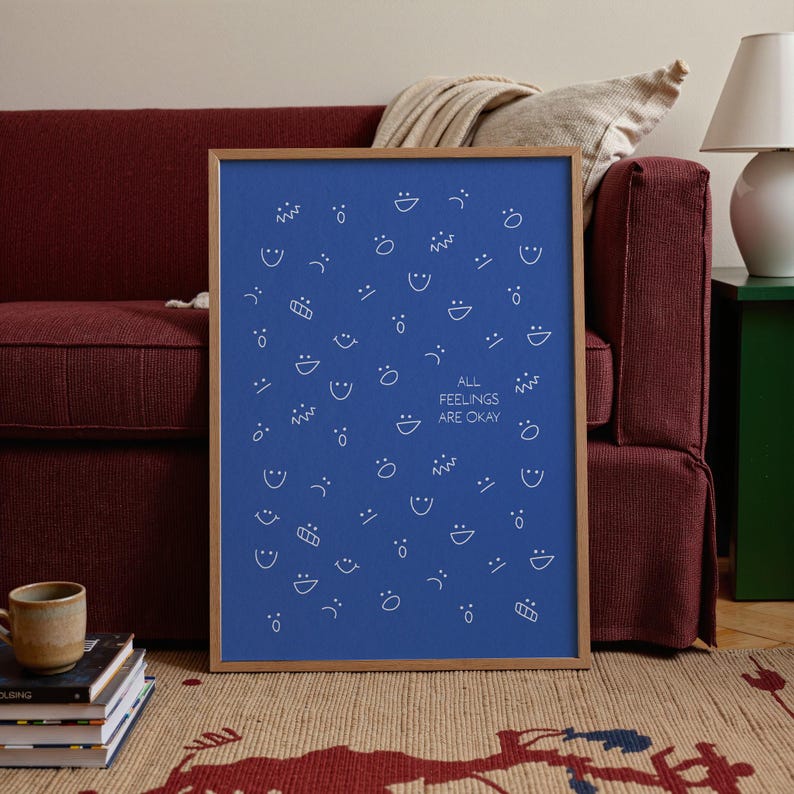 May include: A framed blue print with white line drawings of various facial expressions. The text "ALL FEELINGS ARE OKAY" is centered on the print. The frame is a light brown colour. The print is displayed on a red sofa.
