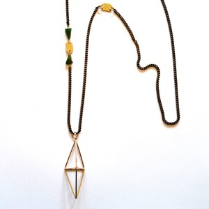 May include: A long necklace with a geometric pendant. The necklace features a dark chain with a gold-colored diamond-shaped pendant. It also has green and gold-colored accents along the chain. The necklace is displayed on a white background.