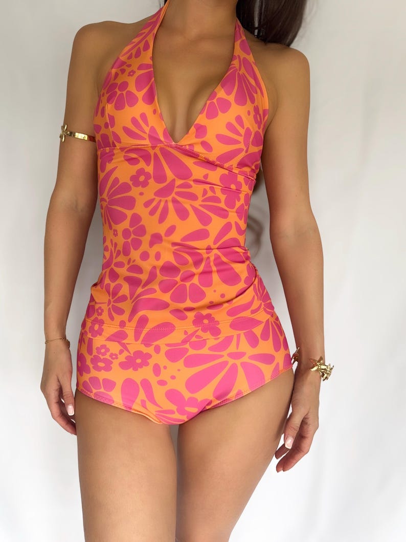 May include: A halter-neck swimsuit in a vibrant orange and pink floral pattern. The one-piece design features a plunging neckline and high-waisted bottoms. The swimsuit is a retro-inspired design.