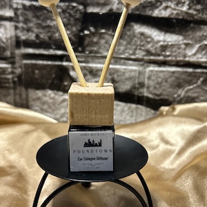 以下が含まれることがあります： 白いボールの付いた木製の棒が2本ついた、小さな黒いボトル。ボトルには「Poundtown Car Cologne Diffuser」と書かれたラベルが付いています。ボトルは黒い金属製のスタンドの上に置かれています。