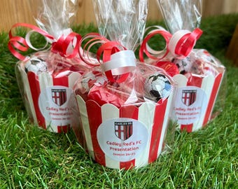Könnte beinhalten: Drei rot-weiß gestreifte Papierbecher gefüllt mit Süßigkeiten und einem Fußball, verpackt in durchsichtigem Zellophan und mit rot-weißen Bändern gebunden. Jeder Becher hat einen Aufkleber mit der Aufschrift "Cadley Red's FC Presentation Saison 24-25."