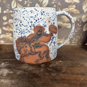 May include: A ceramic mug with a white base and blue speckles featuring a brown, embossed graphic of a cowboy on horseback swinging a lasso. The mug has a cylindrical body and a curved handle on the right side, showcasing a textured finish.