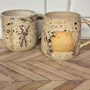May include: Two speckled ceramic mugs with a cream-colored base and dark brown flecks. One mug features a stamped lavender sprig and a small heart. The other mug displays a rectangular plaque with the embossed text "PLANT LAVENDER FOR LUCK".