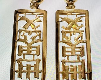 May include: A pair of gold-toned clip-on earrings featuring a rectangular frame with an openwork design. Each earring displays a vertical arrangement of stylized characters within the border, topped with a polished gold-toned spherical stud.