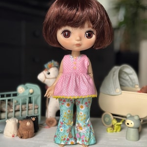 May include: A doll with brown hair and large eyes, dressed in a pink polka-dot top with yellow trim and floral print trousers. The doll is positioned in front of a blurred background with toy accessories.