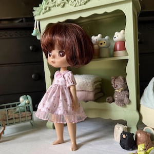 May include: A doll with brown hair and large eyes wearing a pink floral dress with a lace trim. The doll stands in front of a green shelf with decorative bear figurines and a small crocheted teddy bear. A miniature crib is in the background.