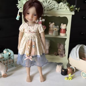 May include: A doll wearing a cream-colored dress with a blue skirt and a pink bow. The doll is standing in front of a green shelf with small bear figurines. A toy horse, a baby carriage, and other toys are also present.