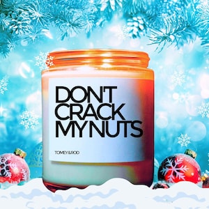 Puede incluir: Un tarro de vela blanco con una etiqueta negra que dice "Don't Crack My Nuts" y el nombre de la marca "Tomey & Rod". El tarro de vela está sentado sobre una superficie blanca con adornos navideños rojos y blancos en el fondo.