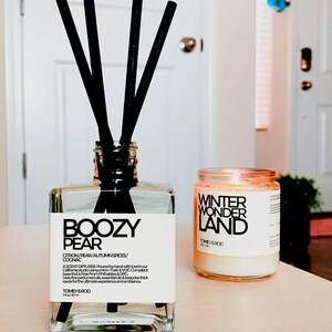 Może przedstawiać: Szklany dyfuzor z przezroczystego szkła z czarnymi trzcinami i białą etykietą z napisem "Boozy Pear Citron/Pear/Autumn Spices/Cognac Ascend Diffuser Poured by hand with love in our California studio using a Non-Toxic & VOC Compliant base that is Free From Phthalates & DPG Uses fine perfumed oils, essential oils & bespoke thick reeds for the ultimate experience and ambiance Tomey & Rod"  Obok dyfuzora znajduje się biała świeca z czarną etykietą z napisem "Winter Wonder Land Tomey & Rod"