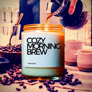 Op de afbeelding: Een witte kaars met een zwart label waarop "Cozy Morning Brew" van "Tomey & Rod" staat. De kaars staat in een glazen pot met een gouden deksel. De kaars is omringd door koffiebonen en er is een koffiekan die koffie in een mok giet.
