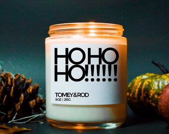 May include: A white candle with a black label that reads "HO HO HO!!!" and "TOMEY & ROD 9 oz | 256g". The candle is lit and glowing.