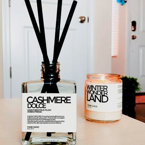 Może przedstawiać: Szklana butelka dyfuzora z czarnymi trzcinami i tekstem "CASHMERE DOLCE" i "DARK FOREST FRUIT/PLUM/AMBER/FREESIA". Butelka stoi na jasnym drewnianym blacie obok przezroczystej szklanej świecy z tekstem "WINTER WONDER LAND".