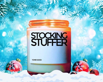 May include: A white candle with an orange lid and a white label that says "Stocking Stuffer" and "Tomey & Rod". The candle is sitting on a white surface with red and white Christmas ornaments.