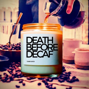 Op de afbeelding: Een kaars met de tekst "DEATH BEFORE DECAF" op een wit etiket. De kaars staat naast een doorzichtige glazen mok, koffiebonen en kleine roze keramische bekers. Koffie wordt uit een karaf gegoten.