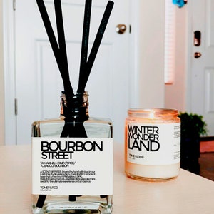 Może przedstawiać: Szklana butelka dyfuzora z czarnymi trzcinami i czarno-białą etykietą z napisem "Bourbon Street" z opisem zapachu "Tamarind/Honey/Spice/Tobacco/Bourbon". Butelka stoi na jasnym drewnianym blacie obok przezroczystej szklanej świecy z białą etykietą z napisem "Winter Wonder Land".