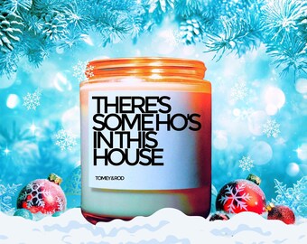 May include: A white candle with a black label that reads "THERE'S SOMEHO'S IN THIS HOUSE" and the brand name "TOMEY & ROD". The candle is sitting on a white surface with red and white Christmas ornaments.