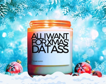 May include: A white candle with a black label that reads "ALL I WANT FOR XMAS IS DAT ASS" and the text "TOMEY & ROD" in smaller font. The candle is sitting on a white surface with red and white Christmas ornaments and a blue and white snowy background.