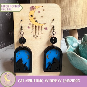 May include: A pair of wooden cat nighttime window earrings. The earrings are black with a blue background and feature a silhouette of a cat looking out a window at a crescent moon and stars. The earrings are displayed on a wooden stand with a moon and stars design.