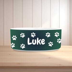 May include: A green and white pet bowl with the name "Luke" in white lettering. The bowl is decorated with white paw prints. The bowl is sitting on a wooden surface.