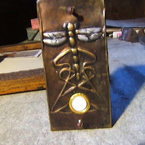 May include: A brass door knocker with a dragonfly design and a white button. The dragonfly is made of copper and has a gold finish.